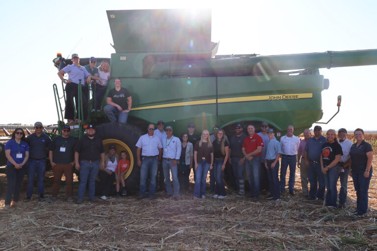 Roughly 28 people — NASA and NASA Acres tour members, Northwestern faculty and students, area producers and their families, including two children — stand together in front of a green John Deere combine in a harvested milo field at the close of the day, backlit by late afternoon sun.