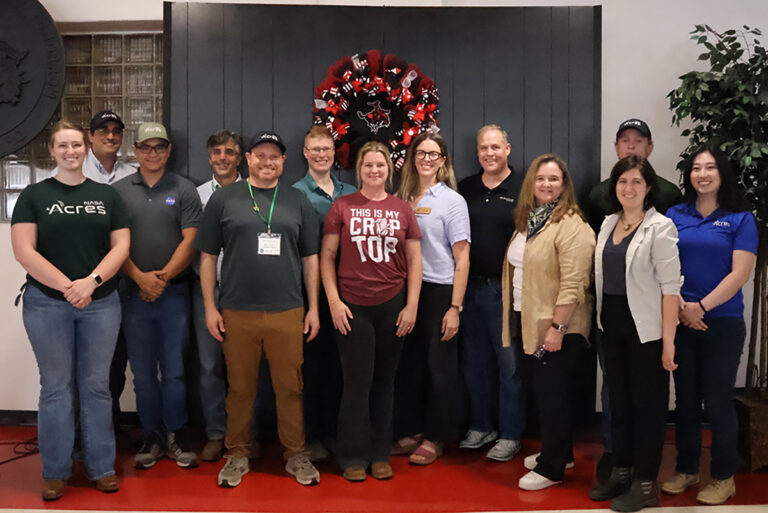 Thirteen NASA and NASA Acres tour members stand together in the Student Center Ballroom at Northwestern Oklahoma State University in front of a red and black wreath bearing the Northwestern Ranger logo. Most wear NASA or NASA Acres branded shirts, polos and caps.