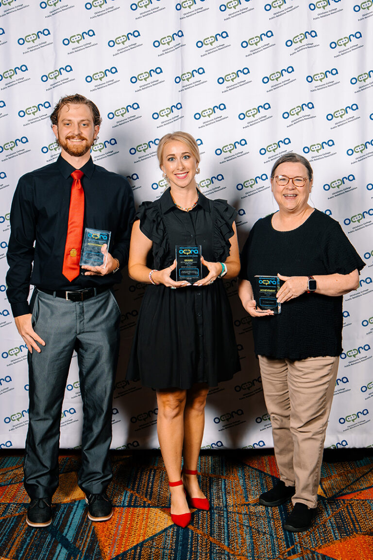 Sean Doherty, Kelsey Martin and Valarie Case of Northwestern Oklahoma State University's University Relations department, each holding an acrylic award, stand in front of an Oklahoma College Public Relations Association backdrop at the 2026 OCPRA awards banquet at Choctaw Casino and Resort in Durant.