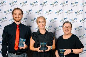 Sean Doherty, Kelsey Martin and Valarie Case of Northwestern Oklahoma State University's University Relations department, each holding an acrylic award, stand in front of an Oklahoma College Public Relations Association backdrop at the 2026 OCPRA awards banquet at Choctaw Casino and Resort in Durant.