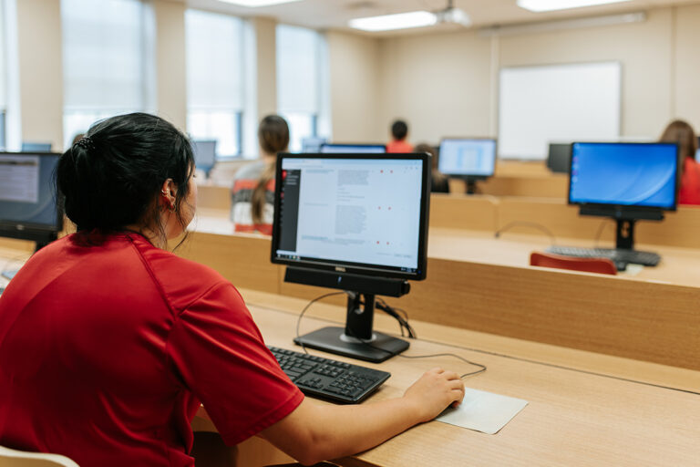 A student in a red shirt works at a desktop computer in a classroom computer lab, with several other students visible at computer stations in the background.