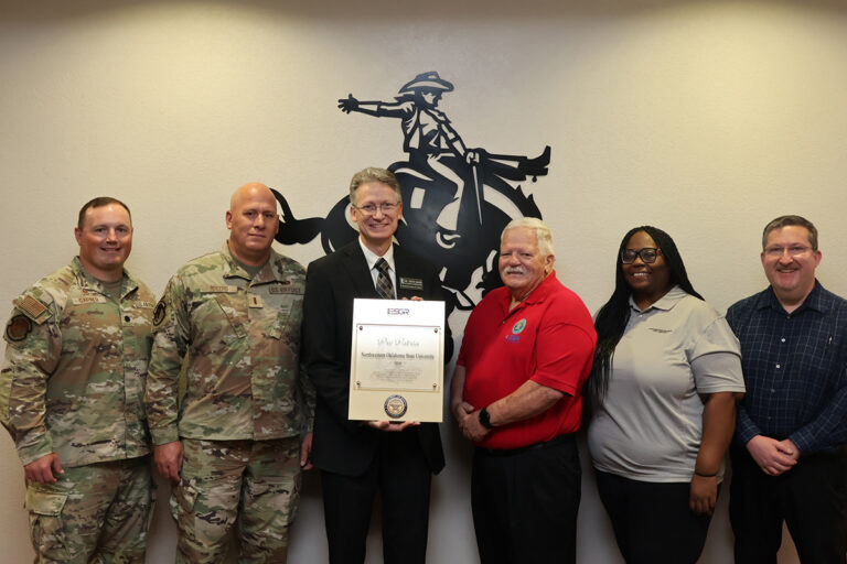 Six people stand in front of a bronc-rider wall emblem in the Student Center Ranger Room; Steve Maier and Paul Ray jointly hold the Pro Patria Award plaque.