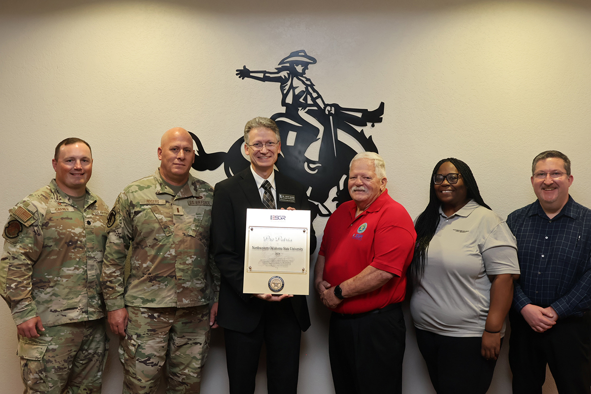 Six people stand in front of a bronc-rider wall emblem in the Student Center Ranger Room; Steve Maier and Paul Ray jointly hold the Pro Patria Award plaque.