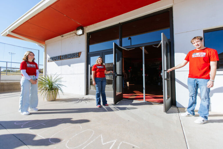 Three Northwestern Oklahoma State University Scholar Ambassadors in red Northwestern T-shirts hold open the glass doors of Percefull Fieldhouse on a sunny day. A sign on the white exterior wall reads "Percefull Fieldhouse," and red and black balloons are visible inside.