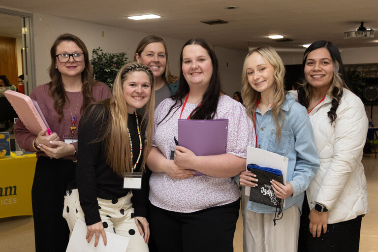 Six Northwestern Oklahoma State University Division of Education students stand together holding folders and notebooks at the Spring 2026 Education Career Fair in the Student Center Ballroom, with employer tables visible behind them.