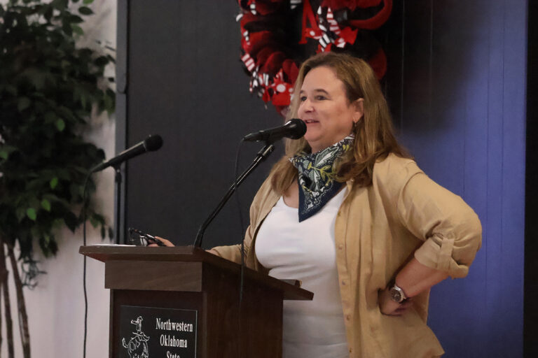 Dr. Karen St. Germain, director of NASA's Earth Science Division, speaks from a podium marked Northwestern Oklahoma State University, wearing a tan jacket and a patterned bandana.