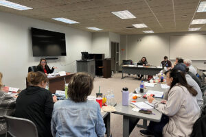 Dr. Donna Devlin, seated behind a desk with a nameplate reading “Donna Devlin,” speaks and gestures to educators seated around rows of gray tables during a Teaching American History seminar at Northwestern Oklahoma State University’s Enid campus.