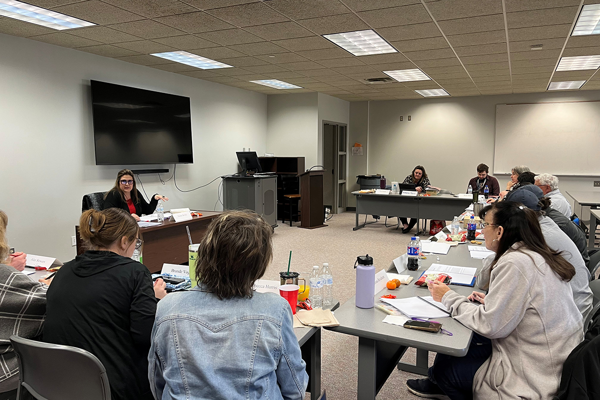 Dr. Donna Devlin, seated behind a desk with a nameplate reading “Donna Devlin,” speaks and gestures to educators seated around rows of gray tables during a Teaching American History seminar at Northwestern Oklahoma State University’s Enid campus.