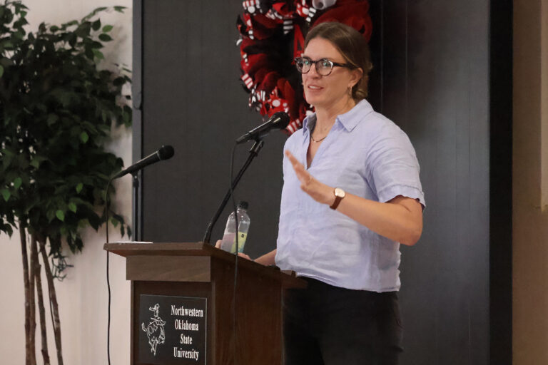 Dr. Alyssa Whitcraft, executive director of NASA Acres, gestures mid-sentence at a podium marked Northwestern Oklahoma State University, wearing a light blue shirt and dark-framed glasses.