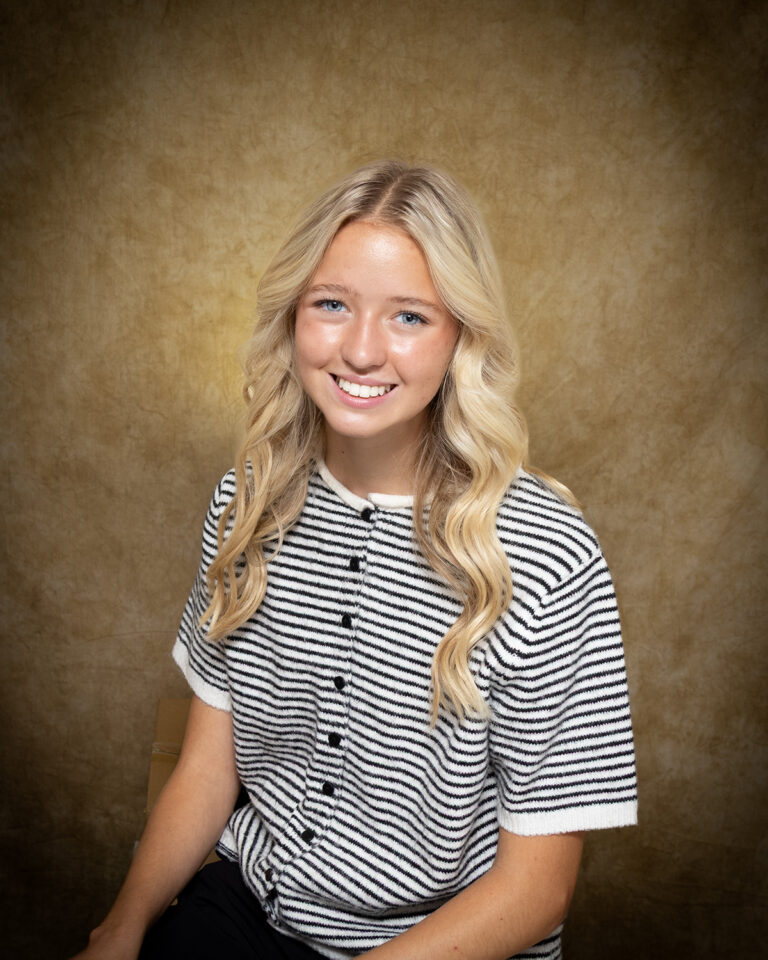 Kinsley Acre, Miss Taloga, one of 17 high school seniors competing in the 74th annual Miss Cinderella Pageant at Northwestern Oklahoma State University, seated for a studio portrait against a mottled tan backdrop, wearing a black and white striped short-sleeve knit top with black buttons, her blond hair worn long and wavy.