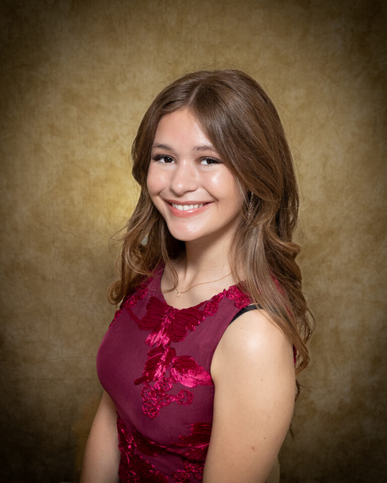Amelie Burgtorf, Miss Shattuck, one of 17 high school seniors competing in the 74th annual Miss Cinderella Pageant at Northwestern Oklahoma State University, seated for a studio portrait against a mottled tan backdrop, wearing a sleeveless burgundy dress with raised floral applique and a thin gold necklace, her light brown hair swept over one shoulder.