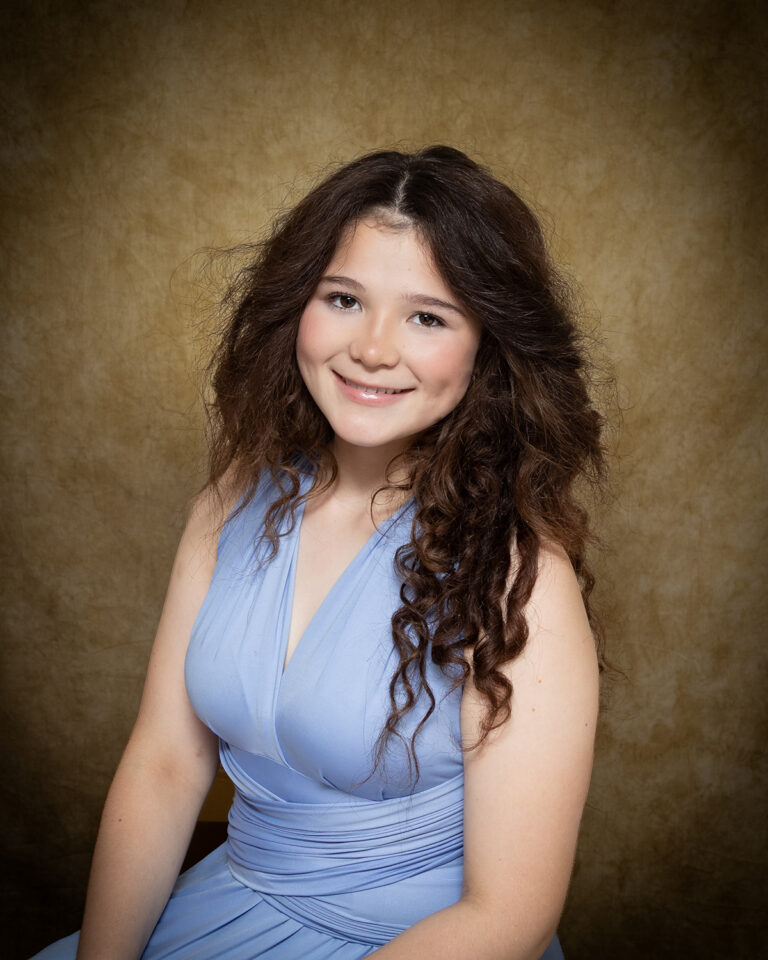 Rebecka Carlson, Miss Burlington, one of 17 high school seniors competing in the 74th annual Miss Cinderella Pageant at Northwestern Oklahoma State University, seated for a studio portrait against a mottled tan backdrop, wearing a light blue sleeveless gown with a gathered waist, her dark brown hair worn long and curly.