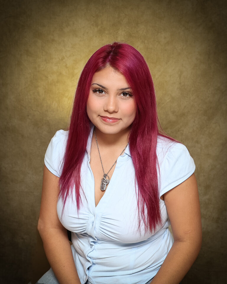 Shai-Ann Nichole Chavez, Miss Timberlake, one of 17 high school seniors competing in the 74th annual Miss Cinderella Pageant at Northwestern Oklahoma State University, seated for a studio portrait against a mottled tan backdrop, wearing a pale blue short-sleeve button-front top and a silver pendant necklace, her magenta hair worn long and straight.