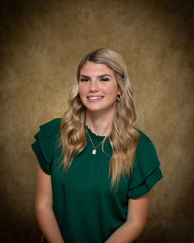 Embrie Crawford, Miss Ringwood, one of 17 high school seniors competing in the 74th annual Miss Cinderella Pageant at Northwestern Oklahoma State University, seated for a studio portrait against a mottled tan backdrop, wearing a dark green top with tiered ruffle sleeves and a gold pendant necklace, her blond hair worn long and wavy.
