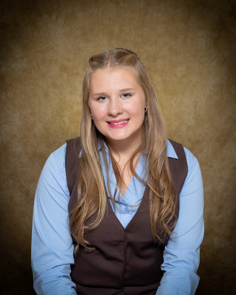 Kaitlyn Culp, Miss Woodward, one of 17 high school seniors competing in the 74th annual Miss Cinderella Pageant at Northwestern Oklahoma State University, seated for a studio portrait against a mottled tan backdrop, wearing a light blue collared shirt under a brown pinstriped vest, her blond hair pulled back at the crown.