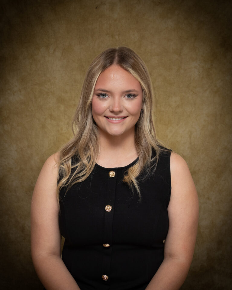 Khloe Cummings, Miss Mooreland, one of 17 high school seniors competing in the 74th annual Miss Cinderella Pageant at Northwestern Oklahoma State University, seated for a studio portrait against a mottled tan backdrop, wearing a black sleeveless dress with gold knot buttons down the front, her blond hair worn long and wavy.