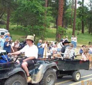 An ATV pulling a small undecorated utility trailer down a parade route, carrying a cooler and a folding lawn chair.
