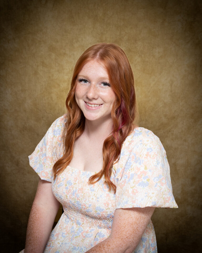 Danica Gleason, Miss Medicine Lodge, one of 17 high school seniors competing in the 74th annual Miss Cinderella Pageant at Northwestern Oklahoma State University, seated for a studio portrait against a mottled tan backdrop, wearing a cream floral-print dress with puffed sleeves, her red hair worn long with a pink strand at one side.
