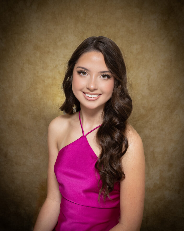 Avyn Harland, Miss Buffalo, one of 17 high school seniors competing in the 74th annual Miss Cinderella Pageant at Northwestern Oklahoma State University, seated for a studio portrait against a mottled tan backdrop, wearing a magenta satin halter dress, her dark brown hair swept over one shoulder.