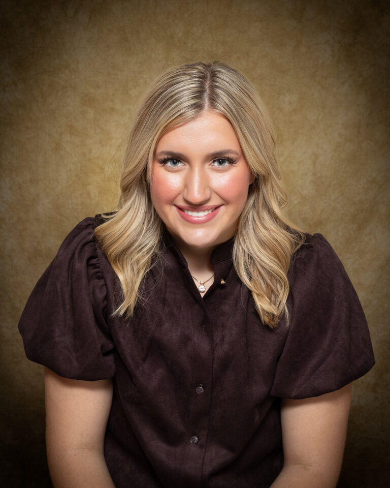 Kassie Irwin, Miss Aline-Cleo, one of 17 high school seniors competing in the 74th annual Miss Cinderella Pageant at Northwestern Oklahoma State University, seated for a studio portrait against a mottled tan backdrop, wearing a dark brown button-front top with puffed sleeves and a pendant necklace, her blond hair worn shoulder length.