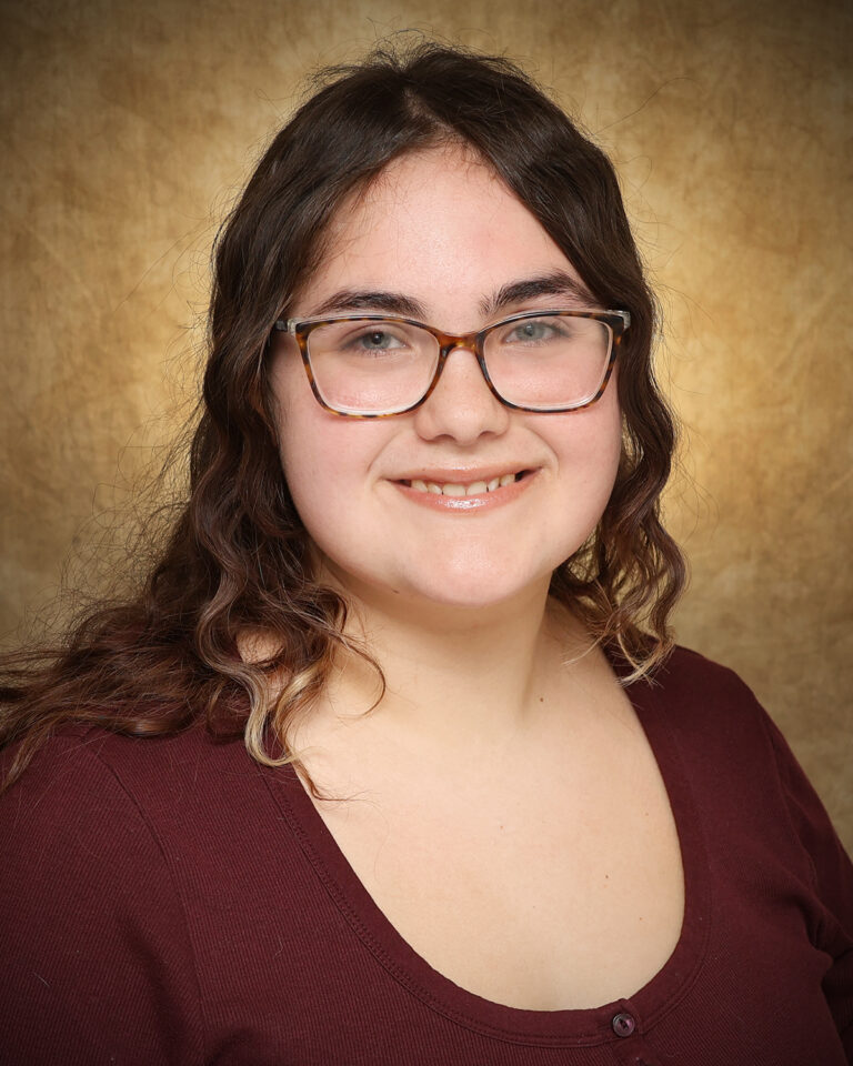 Jacie Willson, a Northwestern Oklahoma State University Upward Bound Math Science student, smiles in a portrait wearing a brown blouse against a mottled gold backdrop.