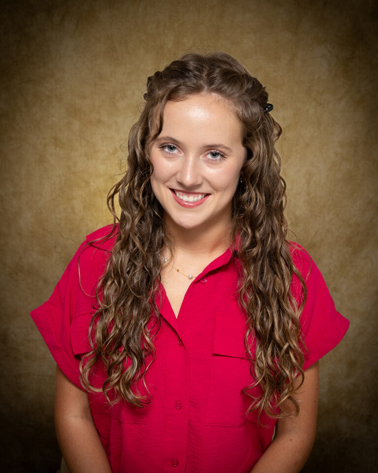 Kylee Logsdon, Miss Alva, one of 17 high school seniors competing in the 74th annual Miss Cinderella Pageant at Northwestern Oklahoma State University, seated for a studio portrait against a mottled tan backdrop, wearing a red short-sleeve button-front shirt and a pearl necklace, her curly hair pulled back at the sides.
