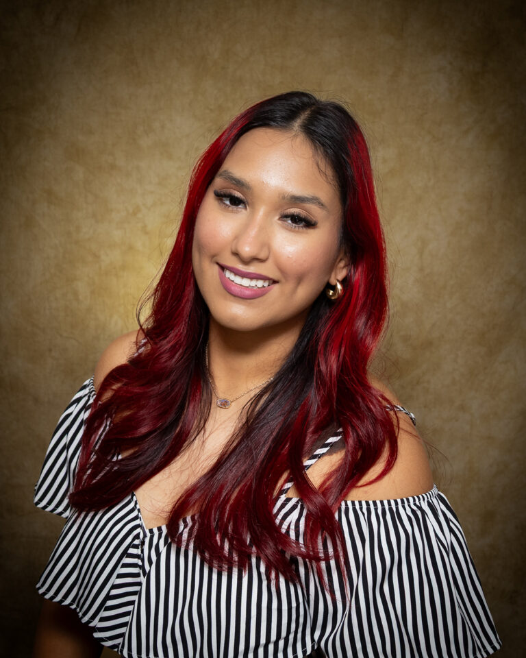 Pricilla Parada, Miss Sharon-Mutual, one of 17 high school seniors competing in the 74th annual Miss Cinderella Pageant at Northwestern Oklahoma State University, seated for a studio portrait against a mottled tan backdrop, wearing a black and white striped off-the-shoulder top and a gold pendant necklace, her hair dyed dark red.