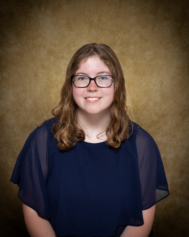 Bristen Robertson, Miss Arnett, one of 17 high school seniors competing in the 74th annual Miss Cinderella Pageant at Northwestern Oklahoma State University, seated for a studio portrait against a mottled tan backdrop, wearing a navy top with sheer flutter sleeves and dark-framed glasses, her brown hair worn shoulder length.