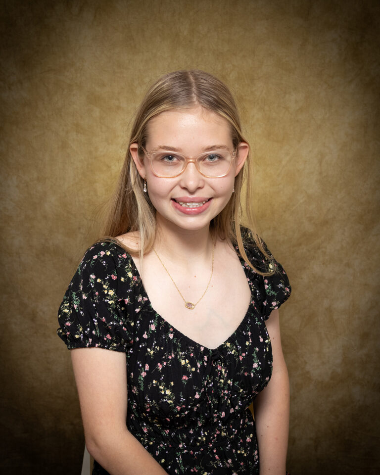 Malinda Sanpaka, Miss South Barber, one of 17 high school seniors competing in the 74th annual Miss Cinderella Pageant at Northwestern Oklahoma State University, seated for a studio portrait against a mottled tan backdrop, wearing a black floral-print dress and clear-framed glasses, her blond hair worn long and straight.