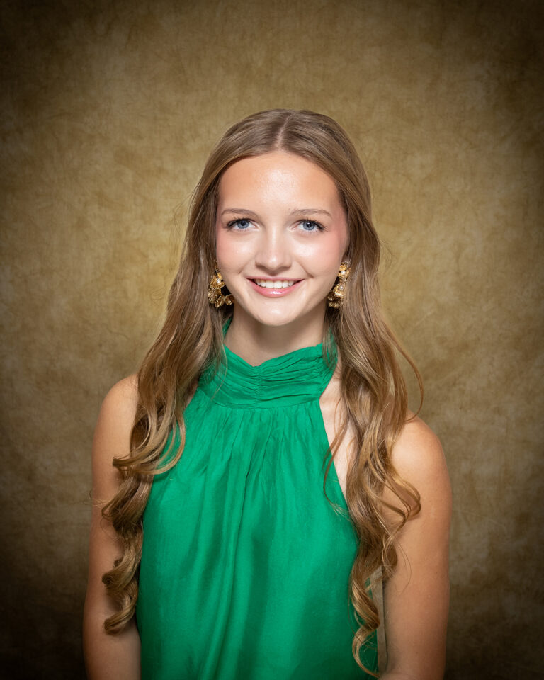 Ava Schultz, Miss Kremlin-Hillsdale, one of 17 high school seniors competing in the 74th annual Miss Cinderella Pageant at Northwestern Oklahoma State University, seated for a studio portrait against a mottled tan backdrop, wearing a green sleeveless halter top and gold statement earrings, her long hair worn loose.