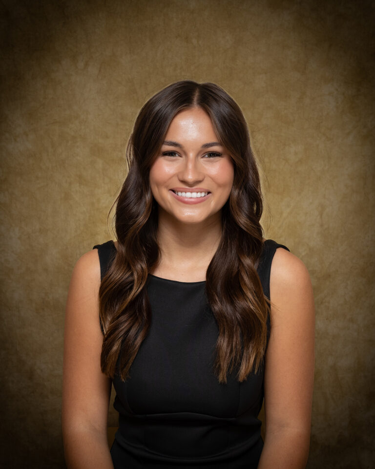 Kaycie Shelton, Miss Cherokee, one of 17 high school seniors competing in the 74th annual Miss Cinderella Pageant at Northwestern Oklahoma State University, seated for a studio portrait against a mottled tan backdrop, wearing a black sleeveless dress, her dark brown hair worn long and wavy.
